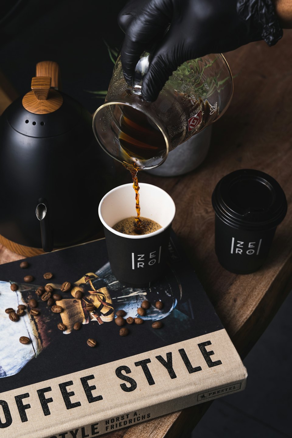 Mit Handschuhen bekleidete Hand gießt Kaffee aus einer Kanne in eine Tasse.